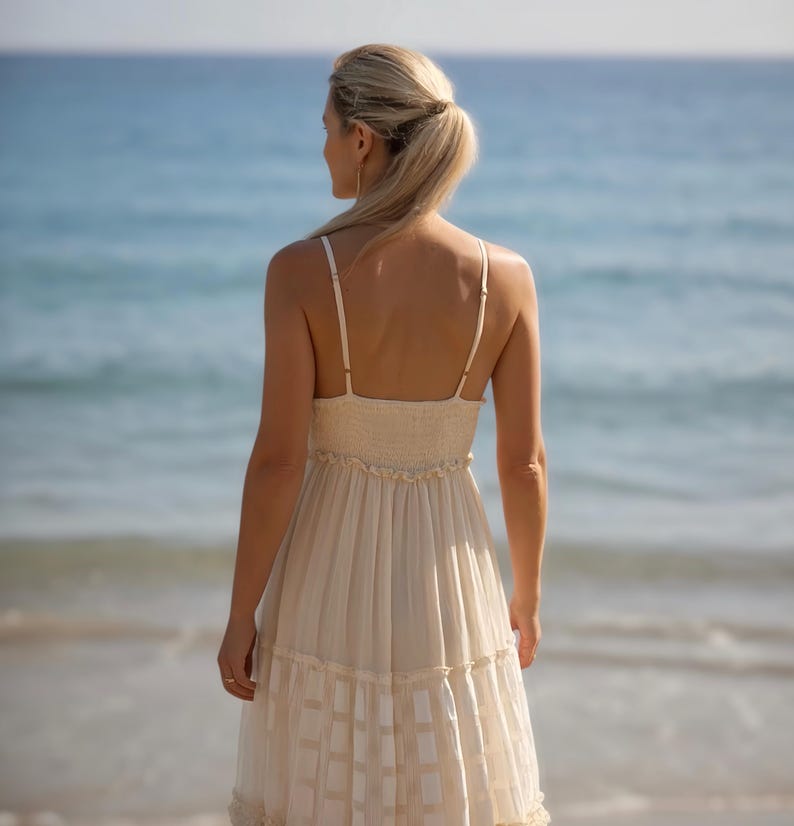 May include: A cream-colored sundress with spaghetti straps and a tiered skirt. The dress has a fitted bodice and a flowing, patterned skirt. The background features a beach and ocean.