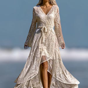 May include: Ivory-colored, long-sleeved dress with a V-neckline and a wrap-style design. The dress features intricate floral lace detailing throughout, a waist tie, and a high-low hemline. The sleeves are long and flared, and the dress has a flowing, ethereal quality.