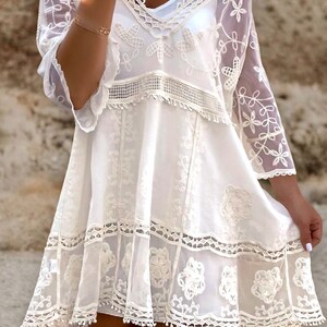 May include: White crochet mini dress with a lace overlay and floral embroidery. The dress has a V-neckline and short sleeves.
