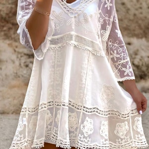May include: White crochet mini dress with a lace overlay and floral embroidery. The dress has a V-neckline and short sleeves.