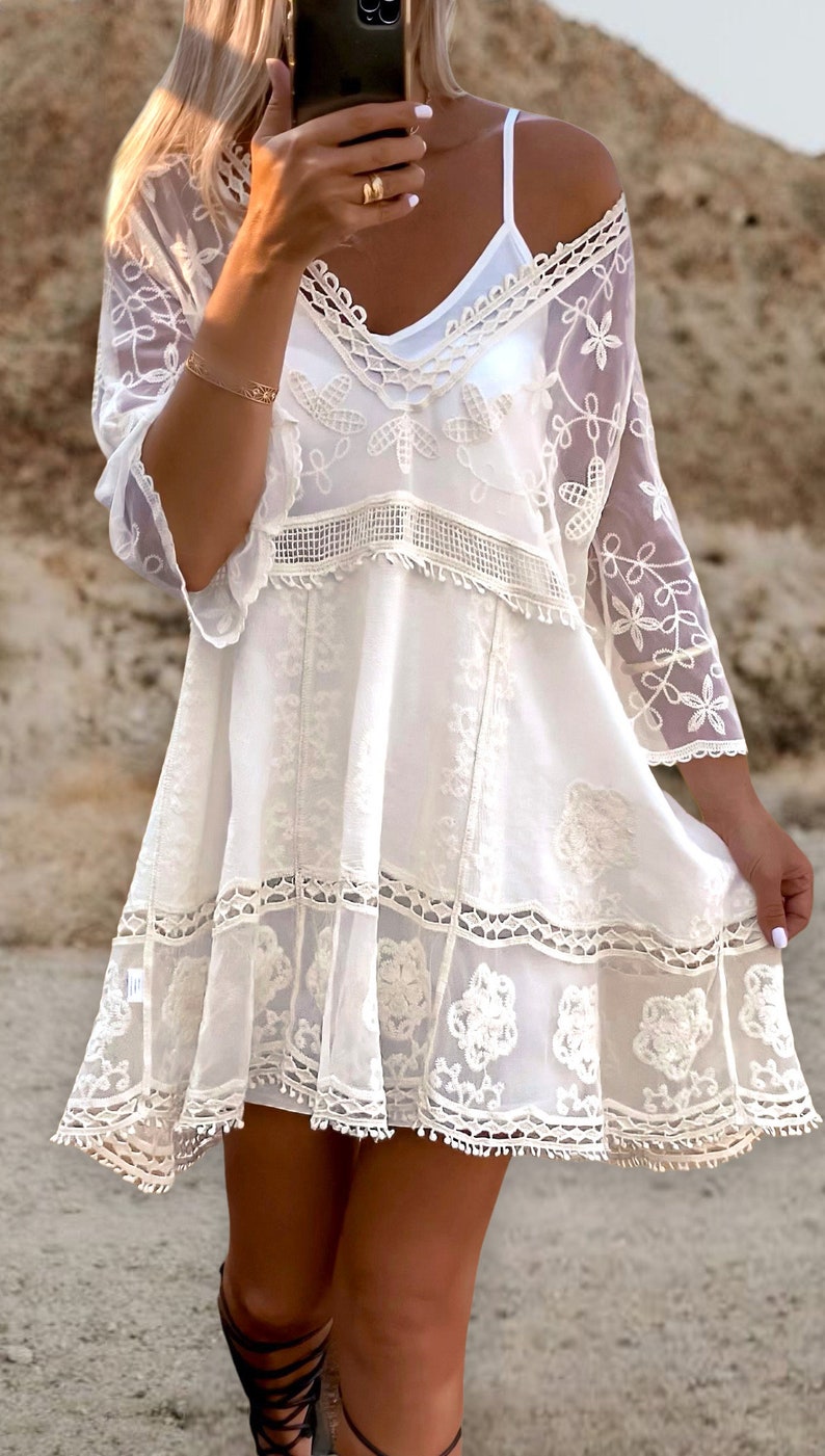 May include: White crochet mini dress with a lace overlay and floral embroidery. The dress has a V-neckline and short sleeves.