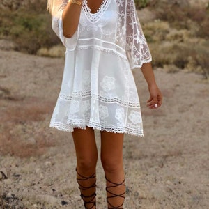 May include: White crochet mini dress with a V-neckline and 3/4 sleeves. The dress has a lace trim and floral embroidery. The model is wearing the dress with brown lace-up sandals.