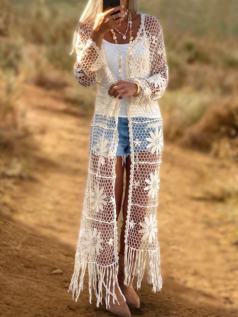 Puede incluir: Un c&aacute;rdigan de crochet blanco con un patr&oacute;n floral y ribete de flecos. El c&aacute;rdigan est&aacute; abierto en la parte delantera y tiene mangas largas.