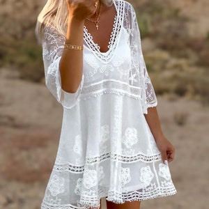 May include: White crochet mini dress with a V-neckline, 3/4 sleeves, and a tiered skirt. The dress features delicate floral embroidery and a lace trim.