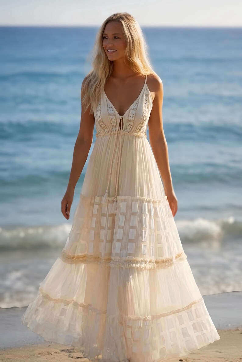 May include: A cream-colored maxi dress with spaghetti straps and a V-neckline. The dress features a tiered skirt with a textured pattern and lace trim. The model is standing on a sandy beach with the ocean in the background.