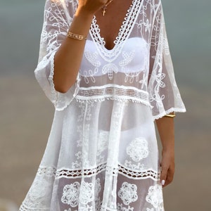 May include: White crochet beach cover-up with a V-neckline, 3/4 sleeves, and a tiered skirt. The cover-up has a delicate floral pattern and a lace trim.