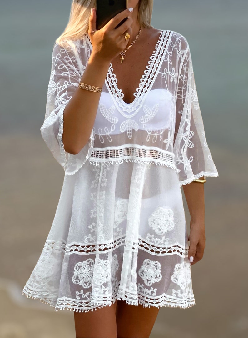 May include: White crochet beach cover-up with a V-neckline, 3/4 sleeves, and a tiered skirt. The cover-up has a delicate floral pattern and a lace trim.