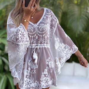 May include: White crochet cover-up with a delicate floral pattern and a tie at the waist. The cover-up has a sheer, lightweight fabric and bell sleeves.