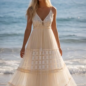May include: A cream-colored maxi dress with spaghetti straps and a V-neckline. The dress features intricate lace detailing on the bodice and a tiered skirt with a geometric pattern. The model is standing on a beach with the ocean in the background.