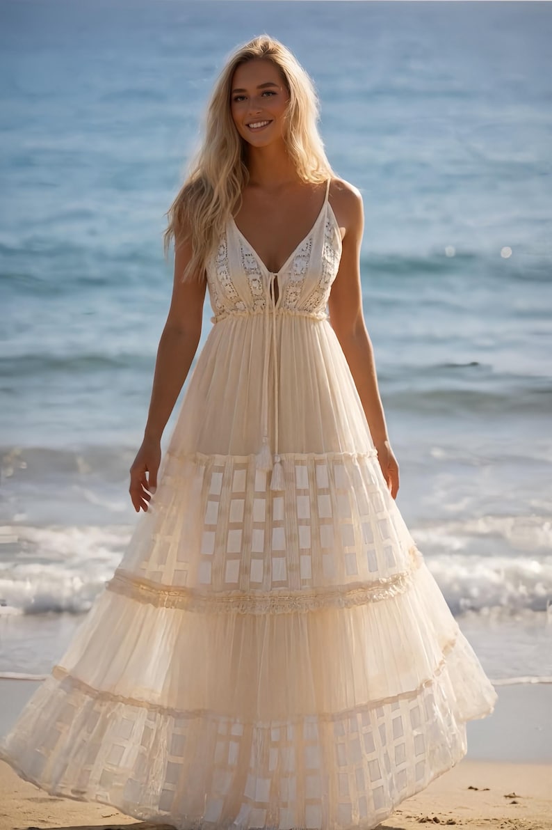 May include: A cream-colored maxi dress with spaghetti straps and a V-neckline. The dress features intricate lace detailing on the bodice and a tiered skirt with a geometric pattern. The model is standing on a beach with the ocean in the background.