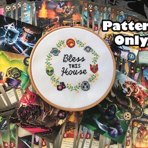 May include: A white embroidery hoop with a cross-stitch pattern that reads "Bless This House". The pattern features a wreath of green leaves with various symbols, including a red heart, a green alien, a blue eye, and a red star. The hoop is surrounded by a variety of trading cards.