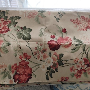May include: Folded fabric with a floral design. The pattern showcases large, open blooms in pink and burgundy, with smaller white flowers and green leaves on a beige backdrop. The fabric is likely cotton or linen, suitable for various home decor projects.