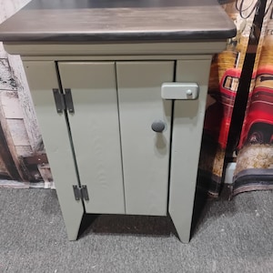 May include: A small, sage green wooden cabinet with a dark brown top. The cabinet has two doors, a grey knob, and a latch. The hinges are dark metal. The cabinet has a rustic, farmhouse style.