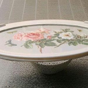May include: An oval serving tray with a floral design featuring pink and white roses, green leaves, and a butterfly. The tray has a cream-coloured rim with handles and a decorative base. The tray is approximately 46 cm long.
