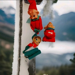 May include: Three vintage Christmas elf figurines in red and green outfits. One elf is skiing down a snowy surface, while the other two stand upright. The background features a snowy landscape and a blurred sky.