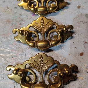 May include: Three antique brass drawer pulls. Each pull features a central wheat stalk design, flanked by curved handles. The pulls have a warm, golden hue and are likely made of solid brass. These decorative hardware pieces are ideal for vintage furniture restoration.