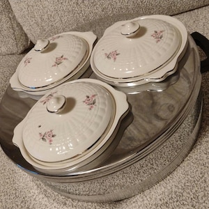 May include: A set of three white porcelain serving dishes with lids, each adorned with a delicate pink floral pattern. The dishes sit on a large, oval, stainless steel serving tray with black handles. The lids have a silver trim and a small, round knob.