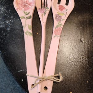 May include: Set of three pink wooden salad utensils tied together with twine. Each utensil features a floral design in shades of pink, purple, and green. The set includes a spoon, a fork, and a slotted spoon.