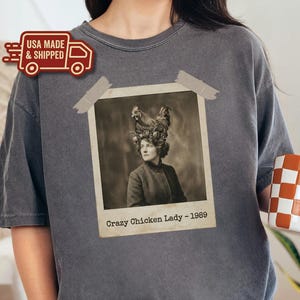 May include: Dark grey t-shirt featuring a vintage photograph of a woman with a chicken headdress. The photo is taped to the shirt and reads "Crazy Chicken Lady - 1989". The shirt has a crew neck and a relaxed fit.