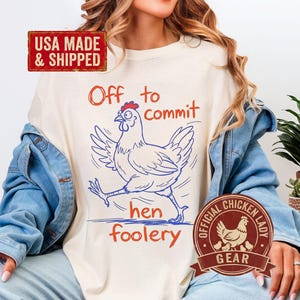 Könnte beinhalten: Cremefarbenes T-Shirt mit einer blau-roten Grafik eines Huhns und den Worten "Off to commit hen foolery". Das Shirt trägt auch den Text "USA MADE & SHIPPED" und "OFFICIAL CHICKEN LADY GEAR."