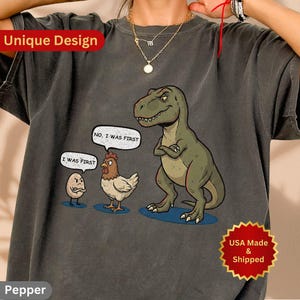 Puede incluir: Camiseta gris oscuro con una ilustración de dibujos animados de un dinosaurio, una gallina y un huevo. El dinosaurio tiene los brazos cruzados, y la gallina y el huevo están discutiendo. Las burbujas de texto dicen "No, yo fui primero" y "Yo fui primero."