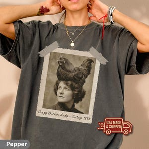 Puede incluir: Camiseta de color pimienta con una imagen en tonos sepia de una mujer con un gallo en la cabeza. La imagen está pegada a la camiseta con cinta gris. El texto de la imagen dice "Crazy Chicken Lady - Vintage 1898".