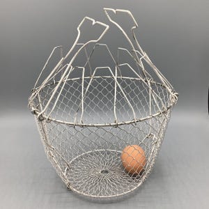 May include: A vintage white wire egg basket with a handle. The basket has a rounded shape with a mesh design and a single brown egg inside. The handle has a unique, decorative shape.