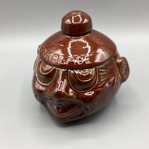 May include: A brown ceramic jar with a lid, featuring a face design with detailed facial features. The jar has the word "ARMALADE" embossed on the lid. The jar is glazed with a glossy finish.