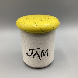May include: A ceramic jam jar with a speckled, off-white body and a bright yellow lid. The lid is dotted with small black specks. The word "JAM" is written in black script on the front of the jar.
