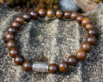 May include: A brown wooden beaded bracelet with a silver metal charm. The bracelet is on a textured gray surface.