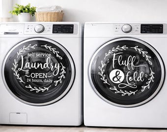 May include: Two white washing machines with black circular doors. One door reads "self service Laundry OPEN 24 hours, daily" in white script. The other door reads "Fluff & Fold" in white script. Both doors have a white floral wreath design.