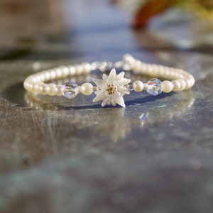 Può includere: Un delicato braccialetto con perle finte bianco crema e un ciondolo centrale a forma di fiore bianco con dettagli dorati. Perline di cristallo trasparente affiancano il fiore. Il braccialetto è infilato su una catena color argento.