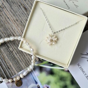 Può includere: Una collana a catena argentata con un ciondolo a forma di fiore con petali bianchi e centro dorato, presentata in una scatola quadrata, insieme a un bracciale di perle d'acqua dolce bianche con un piccolo ciondolo rotondo in metallo dorato.