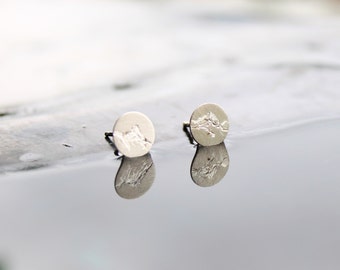 May include: A pair of circular silver stud earrings featuring a textured mountain range design on the front surface. The earrings have a matte finish and are positioned upright, showing the post backing and a clear reflection on the surface.