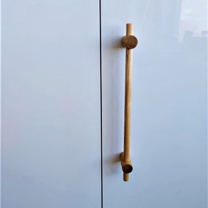 Puede incluir: Una manija de puerta de armario de madera con una forma cilíndrica larga y delgada y dos pomos redondos en la parte superior e inferior.