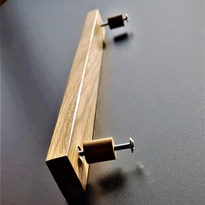 Puede incluir: Un tirador de cajón de madera con forma rectangular y acabado marrón claro. El tirador tiene dos tornillos plateados en la parte superior e inferior.