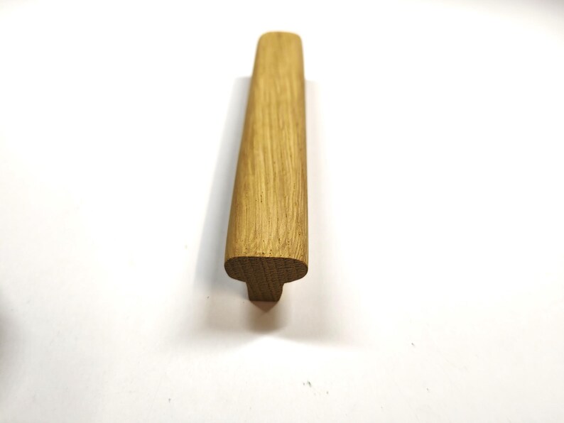 May include: A light brown wooden cabinet handle with a smooth, elongated top surface and a T-shaped profile. The natural wood grain is visible across the length of the handle, which features rounded edges and a solid, sturdy construction.