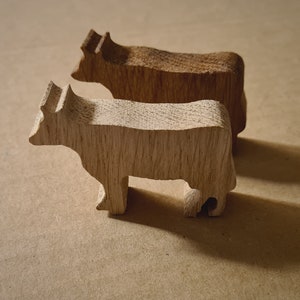 以下が含まれることがあります： 茶色の表面に、薄い茶色と濃い茶色の木製の牛の置物が2つ並んでいます。