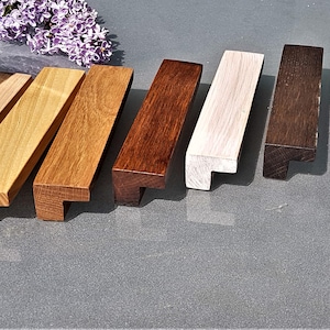 Könnte beinhalten: Sieben Holzknaufgriffe für Schranktüren in verschiedenen Holzbeizen, darunter Natur, Hellbraun, Dunkelbraun und Weiß. Die Griffe sind alle gleich groß und haben die gleiche Form, mit einem quadratischen Sockel und einer abgerundeten Oberseite.