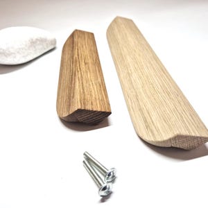 Könnte beinhalten: Zwei Schrankgriffe aus Holz mit abgeschrägter Kante, einer in dunkelbrauner Holzmaserung und einer in heller natürlicher Holzmaserung, begleitet von einem glatten weißen Stein und zwei silbernen Metallschrauben zur Montage an Möbeln.
