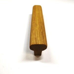 May include: A long, rectangular cabinet handle crafted from natural light brown wood with a visible grain pattern. The handle features a smooth, rounded top surface and a narrower base, presenting a clean, linear profile with a warm, organic finish.