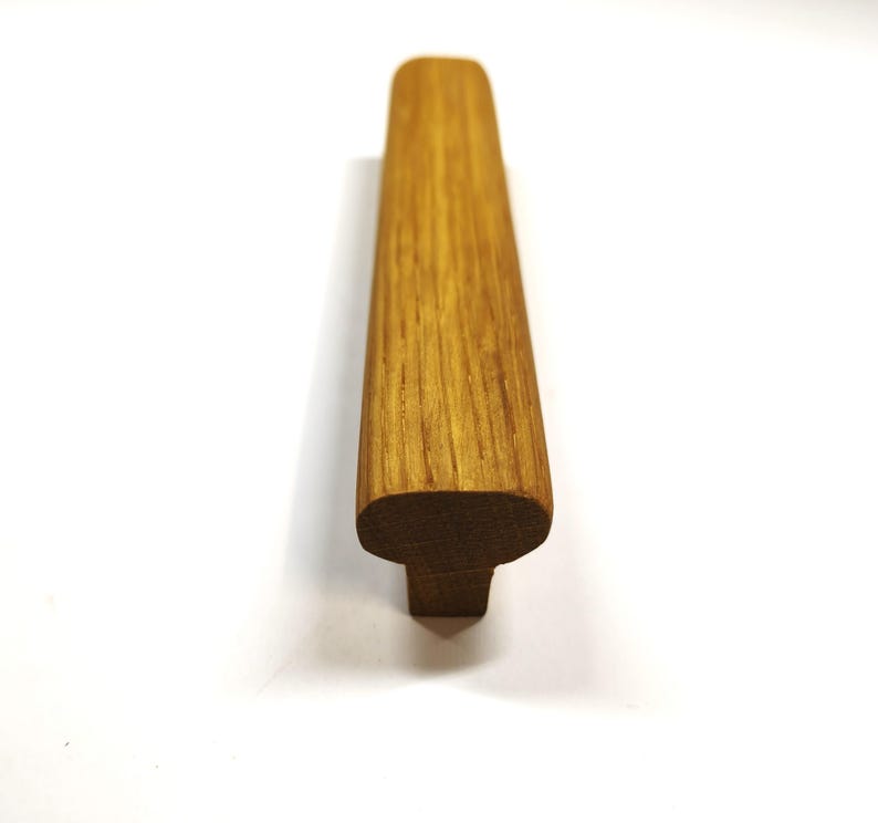 May include: A long, rectangular cabinet handle crafted from natural light brown wood with a visible grain pattern. The handle features a smooth, rounded top surface and a narrower base, presenting a clean, linear profile with a warm, organic finish.
