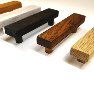 May include: Five rectangular wooden cabinet pulls arranged in a row. The handles feature different finishes including light oak, medium brown, dark brown, black, and a white-washed wood grain, each with two cylindrical mounting posts underneath.