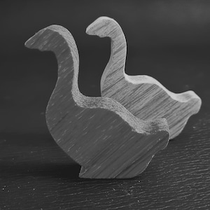 Peut inclure: Deux figurines de canards en bois, sculptées dans un style simple et minimaliste. Les canards sont tournés dans la même direction, celui du fond étant légèrement plus haut que celui du devant.