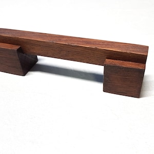 May include: A handcrafted, rectangular wooden drawer pull with a rich, dark brown finish. The pull features a horizontal bar supported by two square blocks, creating a minimalist and modern design. The wood grain is visible.