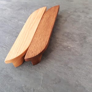 Può includere: Due maniglie per armadi in legno, una in legno naturale e l'altra in legno tinto di marrone scuro. Entrambe hanno la forma di un rettangolo arrotondato con una leggera curva nella parte superiore.