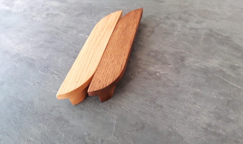 Op de afbeelding: Twee houten kastgrepen, een van naturel hout en een donkerbruin gebeitst. Beide hebben de vorm van een afgeronde rechthoek met een lichte bocht aan de bovenkant.