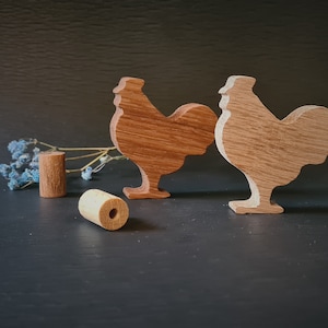 Peut inclure: Deux figurines de coq en bois avec une finition en bois naturel. Les coqs sont debout sur une surface sombre avec un petit bouquet de fleurs bleues séchées et deux chevilles en bois.