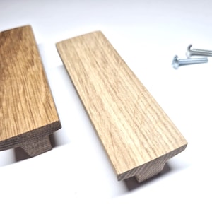 Könnte beinhalten: Zwei rechteckige Schrankgriffe aus Holz mit einer natürlichen hellbraunen Holzmaserung. Die Griffe haben ein T-förmiges Profil mit einer flachen Oberseite und einem zentralen Befestigungssockel. Zwei silberfarbene Metallschrauben sind für die Montage enthalten.