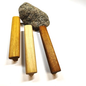 May include: Three elongated wooden cabinet pulls in varying shades of light, medium, and dark brown wood grain, positioned vertically near a single grey speckled stone. Each handle features a T-shaped profile with a flat top and a narrow base.